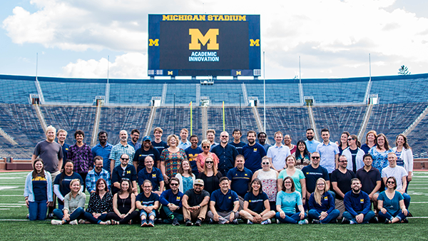 Photo of several individuals posing for the camera in the middle of Michigan Stadium with the Office of Academic Innovation logo on the jumbotron.