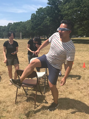 A blindfolded student lifting his leg over a folding chair while two students laugh in the background