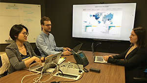 Eejain Huang, Filip Jankovic and Kara Foley in discussion while sitting around a table with laptops in front of large screen with a visualization of the globe