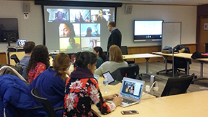 Students seated around a long row of tables with professor Don Peurach standing at the front of room looking at a projection screen with seven individuals on webcams.