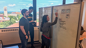 Three staff members collaborating around a white board with a view of campus in the background