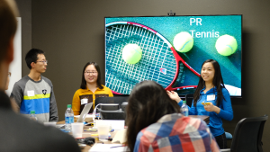 Three students presenting to a large group in front of a screen with an image titled "PR Tennis"