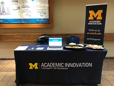 A table covered by an Academic Innovation tablecloth with a laptop on other materials on top and an Academic Innovation banner standing next to the table