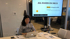 Yuanru Tan seated at the end of a large conference room tab with a monitor behind her reading "All Hands on Deck, Alt Text Writing Jam @DEIL"