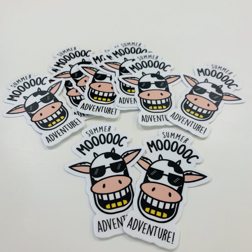 Photo of an assortment of stickers with an illustration of a smiling cow with the words "Summer MOOOOC Adventure."