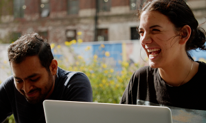 A male student on the left laughing with a female student on the right with her laptop open. 