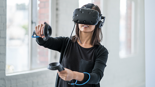 A woman wearing a virtual reality headset and holding two VR devices in her hand with one hand held higher than the other.