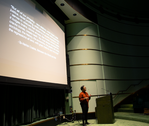 Dr. Courtney Cogburn speaking to an audience and standing in front of a large projection screen in a lecture hall.