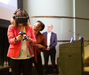 Rachel Niemer facing the camera wearing a VR headset and holding her arms out while Courtney Cogburn and Jeremy Nelson view a large projector screen behind her.
