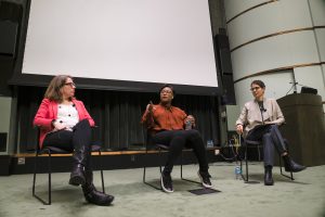 Rachel Niemer, Courtney Cogburn, and professor Sara Blair seated with Courtney Cogburn speaking to an audience in a large lecture hall.