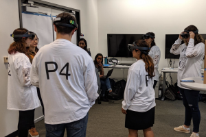 (A group of eight students, five of whom are wearing virtual reality headsets, standing in a circle in a classroom.)