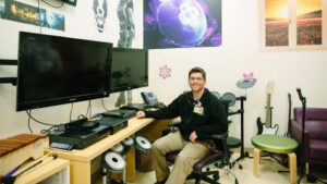 JJ Bouchard at a technology hub desk with video game system monitors, instruments and more