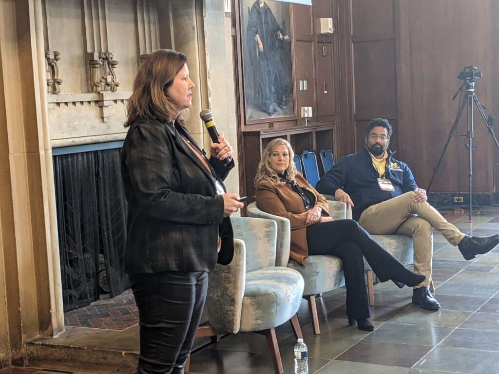 woman standing up holding a microphone while two panelists are seated listening