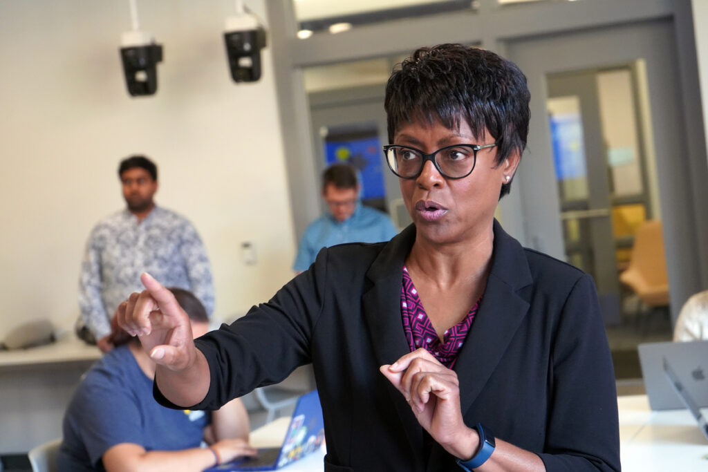 Woman stands and gestures at something inside a large room with various students and staff members sitting and standing behind her.