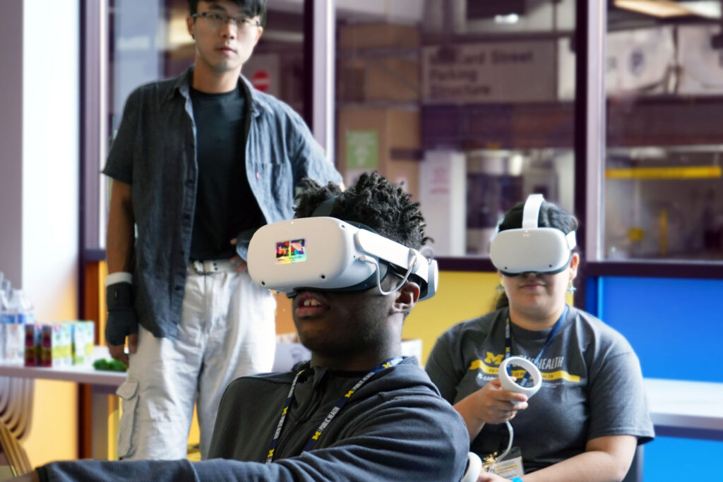 Two students, one in foreground, one in background, sitting with VR headsets and joysticks in their hands. Man in background stands and observes.