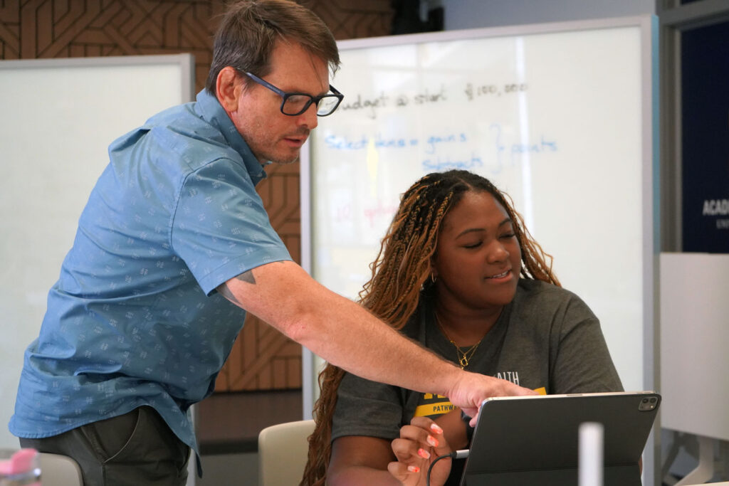 Man, standing, points to something on tablet while student, sitting, looks on. 