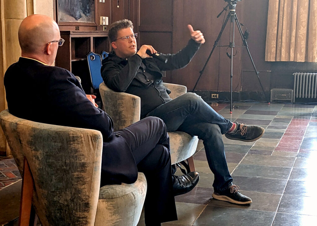 two men holding microphones sitting in chairs having a conversation. 