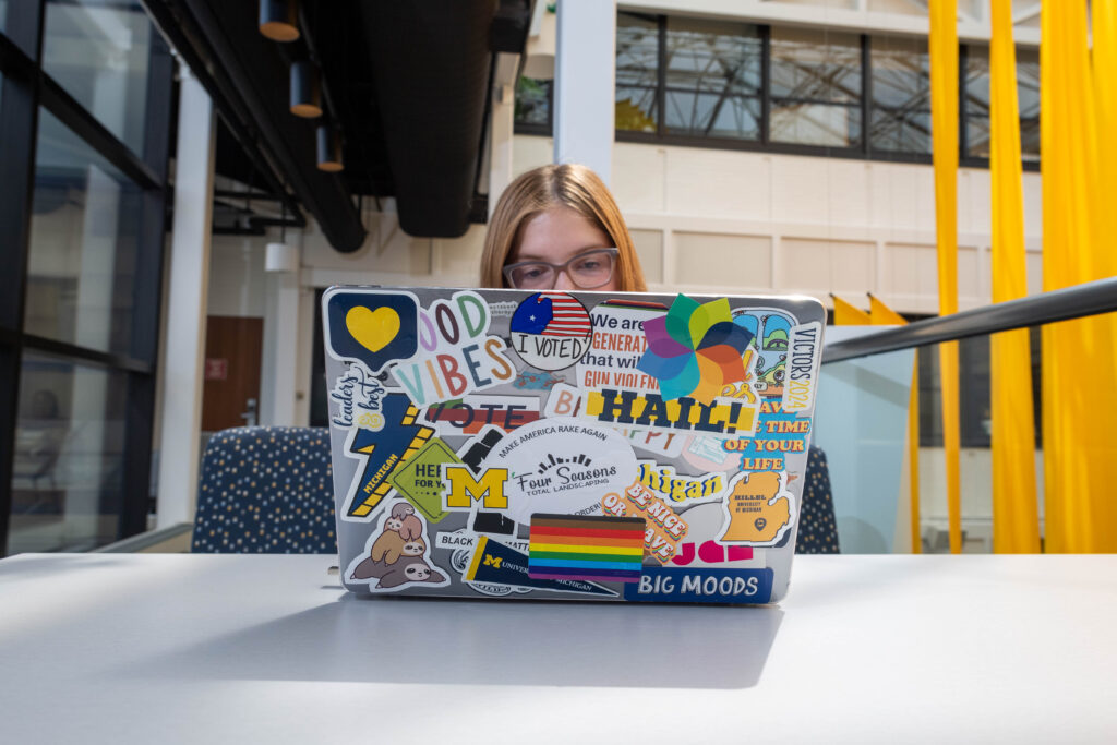 U-M female student sitting at table with laptop in front of her adorned with various stickers and decals. 