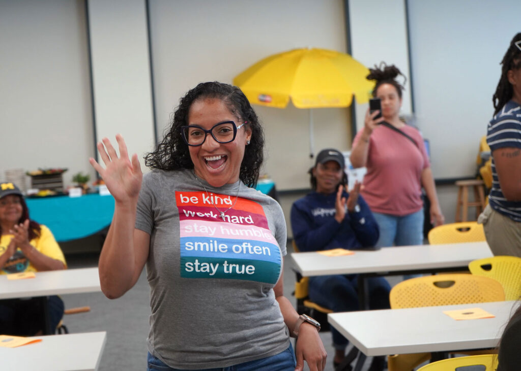 Woman with a t-shirt that says "be kind work hard stay humble smile often stay true" waves to the camera as she walks to the front of the room. 