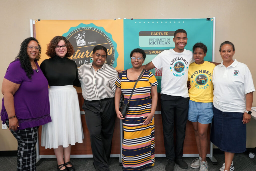 A group poses for a photo in front of a banner for Saturdays in the D. 