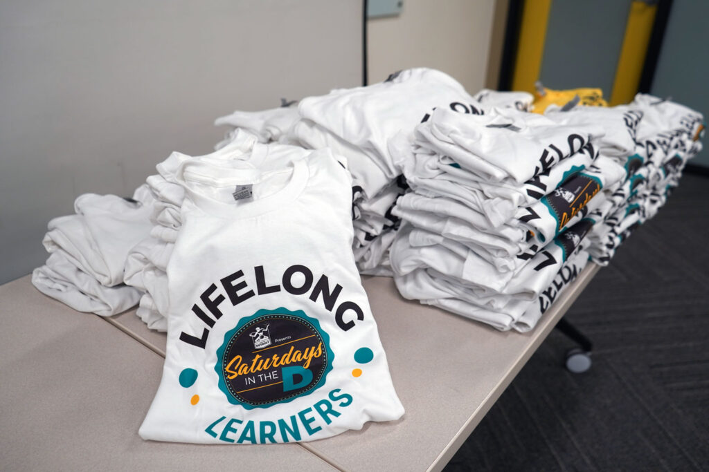 Several piles of shirts, white and yellow, that say "Lifelong Learners" in curved text with the Saturdays in the D logo in the middle. 