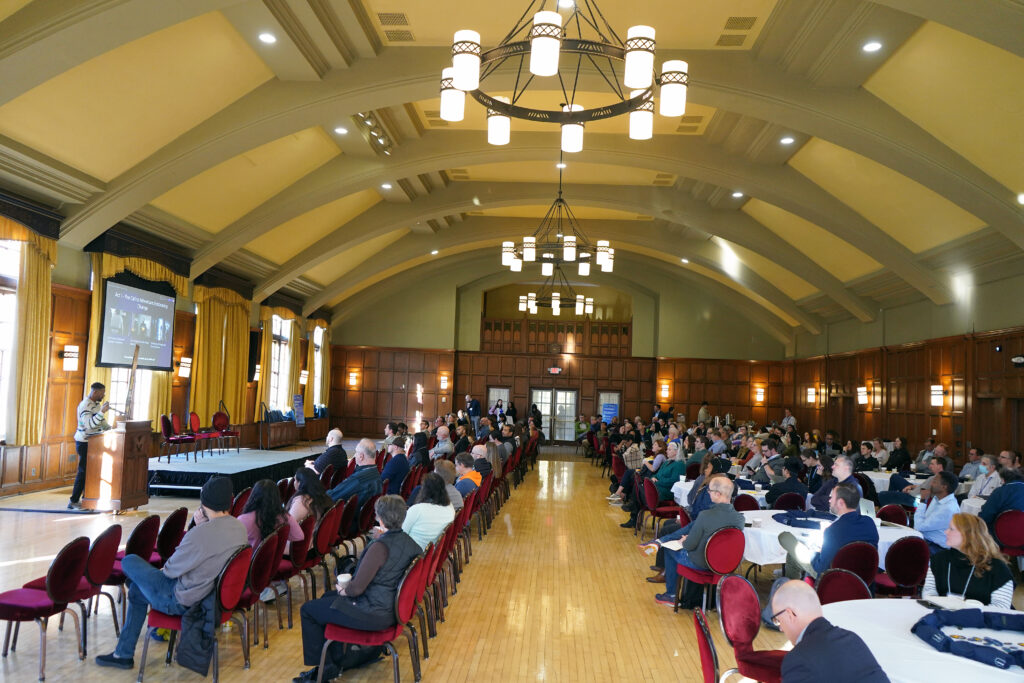 The Michigan League ballroom with people sitting at chairs and at tables listening to a speaker.