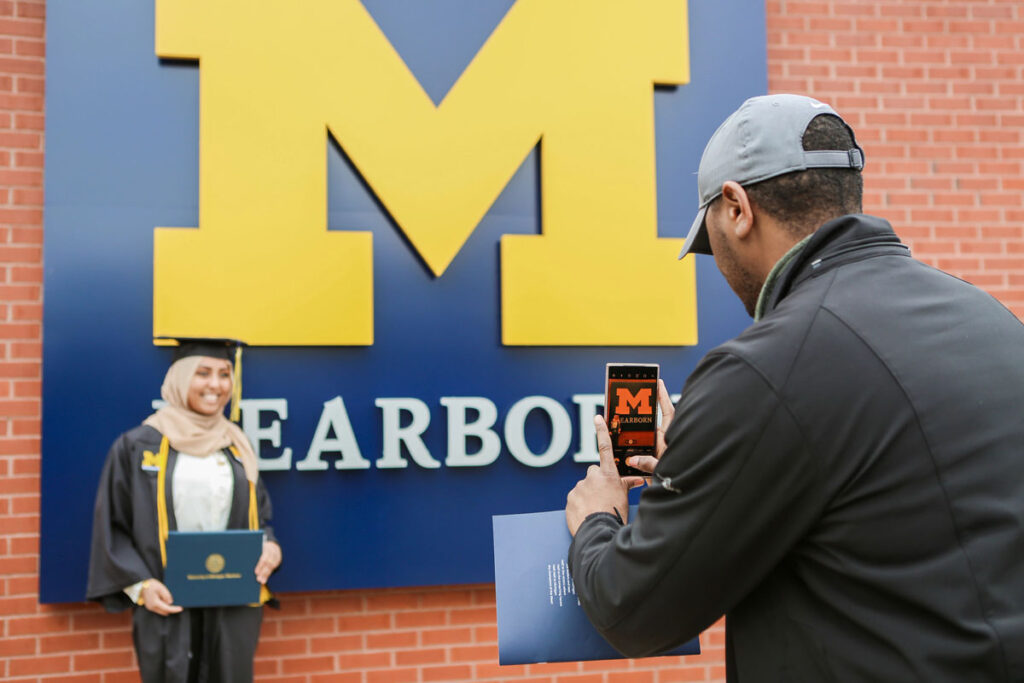 A student wearing a cap and gown poses for a picture while someone takes a photo on a phone.