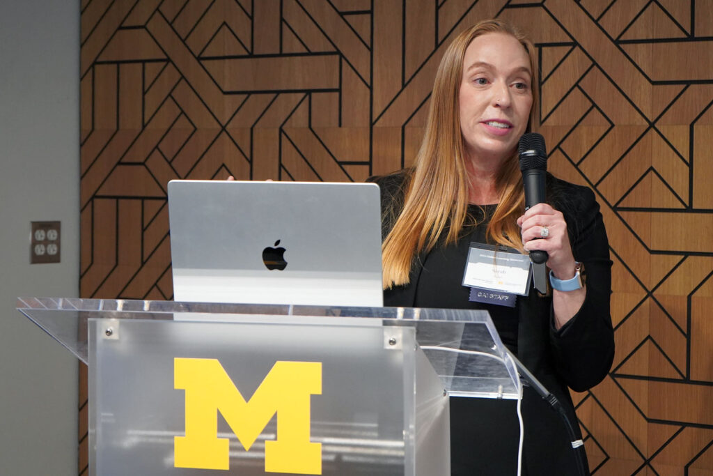 Woman holds a microphone and stands in front of podium that has a block M in front  and a laptop on top. 