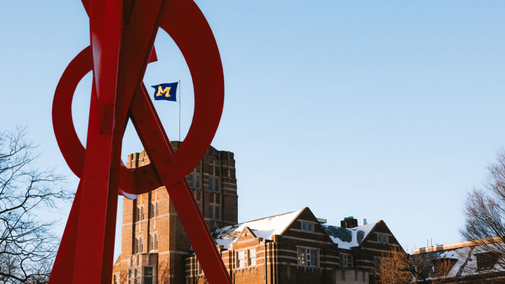 Photo of sculpture with University of Michigan block M flag flying on top of building in background.