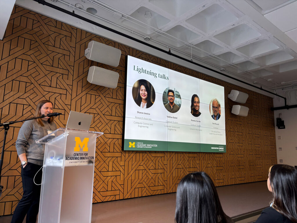 Becky Matz, research scientist lead at the Center for Academic Innovation, stands at a podium with a large display screen featuring headshots of the speakers for the Research & Analytics Showcase at the center.