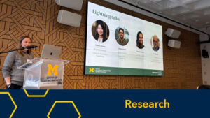 A woman stands in front of a clear podium featuring the University of Michigan Block M logo and Center for Academic Innovation. Behind her is a large display featuring the headshots for four speakers at the Research and Analytics Showcase.