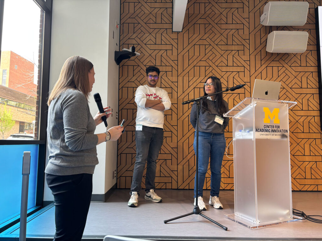 Becky Matz, who is holding a microphone, faces Vaibhav Balloli and Sharon Jessica, who are standing on the stage behind a podium which features the Center for Academic Innovation logo.