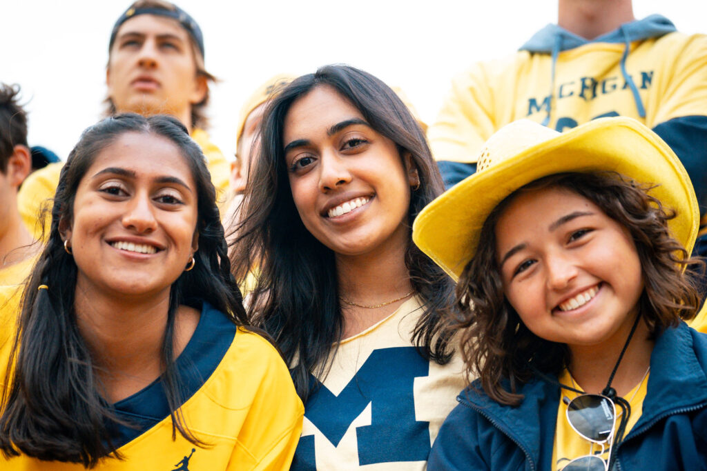 Three students pose for a photo with various pieces of Michigan clothing on, the person in the middle has a big Block M on their T-shirt.