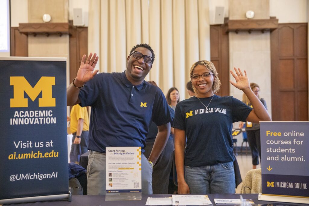 Two people at a table waving straight ahead. A small banner on the table says Academic Innovation with a Block M. A placard on the right says Free online courses for students and alumni with a Michigan Online logo.