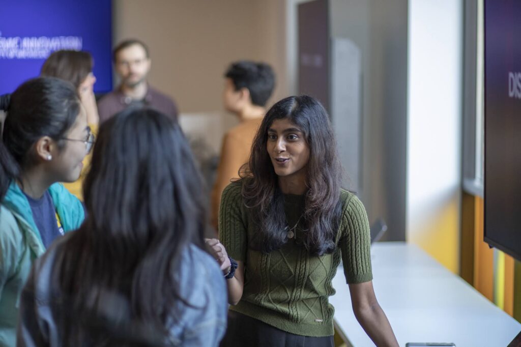 A group chats during an event. A woman is speaking to two others and several people appear talking in the background.