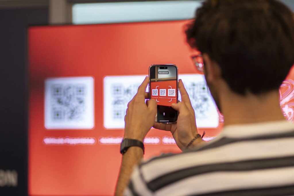 A person holds their phone up to a screen to capture one of several QR codes on the screen.