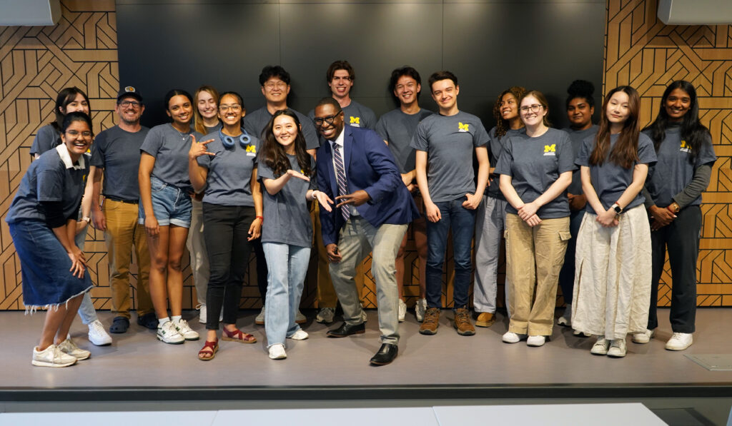 Group of student fellows in matching T-shirts pose for a photo with supervisor in center of photo wearing a blue dress jacket and gray slacks.