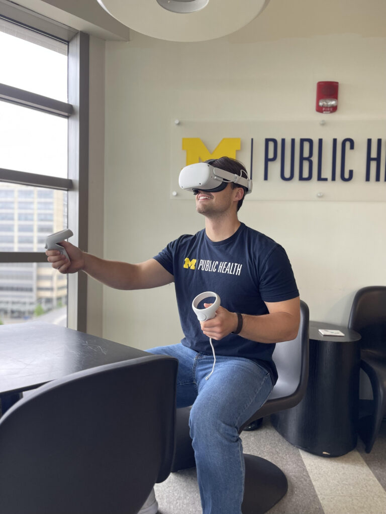 Man sitting in chair with VR headset and a controller in either hand. He is wearing a Michigan School of Public Health T-shirt