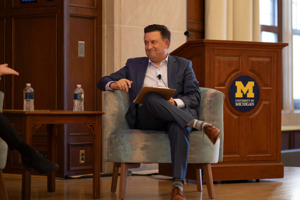 A man in a navy suit smiles while seated with a folder in an armchair on a dais near a University of Michigan podium.