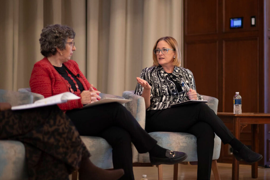 Two women sit on armchairs on a dais. The woman on the right is wearing glasses and gesturing in conversation; the woman on the left in a red sweater is listening with hands crossed on her lap.