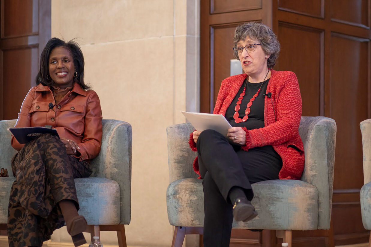 Two women are seated in armchairs on a dais. One smiles while holding notes; another speaks while holding papers.