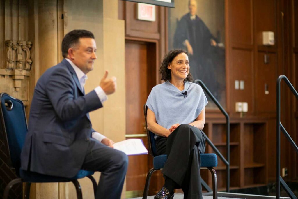 Man and woman sitting and looking out to a crowd out of frame. Man is holding notepad and gesturing while speaking to crowd.