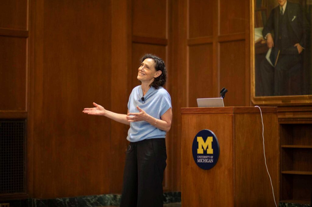 Woman stands in front of podium with a University of Michigan logo and gestures with her hands while smiling. 
