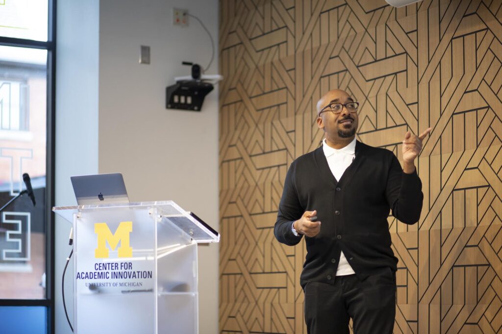 Man standing points to something out of frame. There is a podium next to him with a Center for Academic Innovation logo on the front of the podium, and a laptop on top of podium.