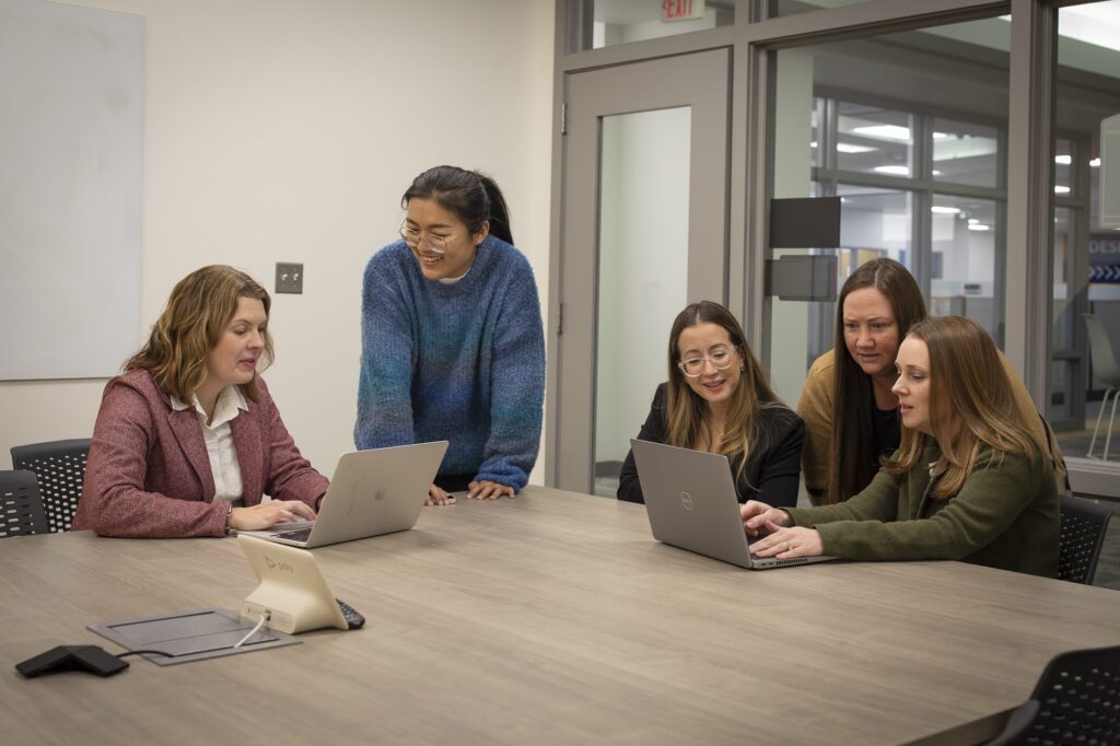 Group of five reviews content on two laptops while working at a large conference table.