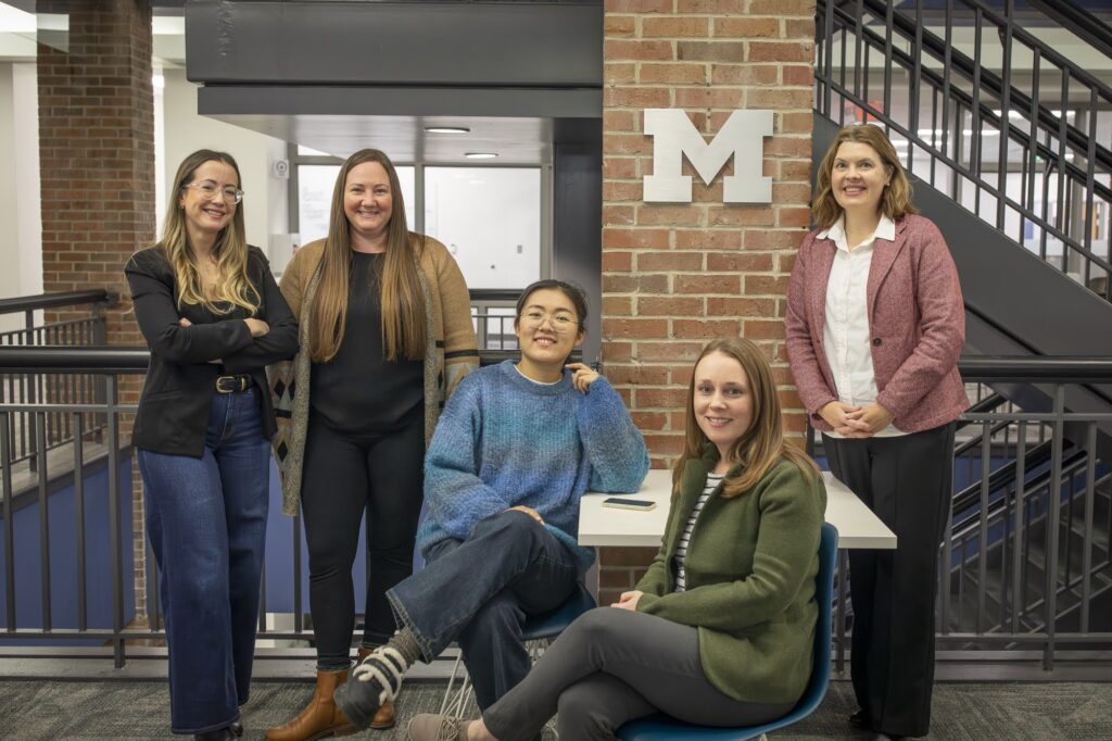 Five women pose for a photo around a table with a brick column that has a silver block M in the background.