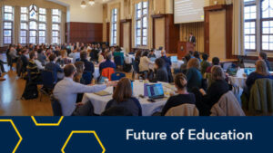 event full of attendees at round tables listening to a speaker on stage at a podium. graphic on bottom says "Future of Education"