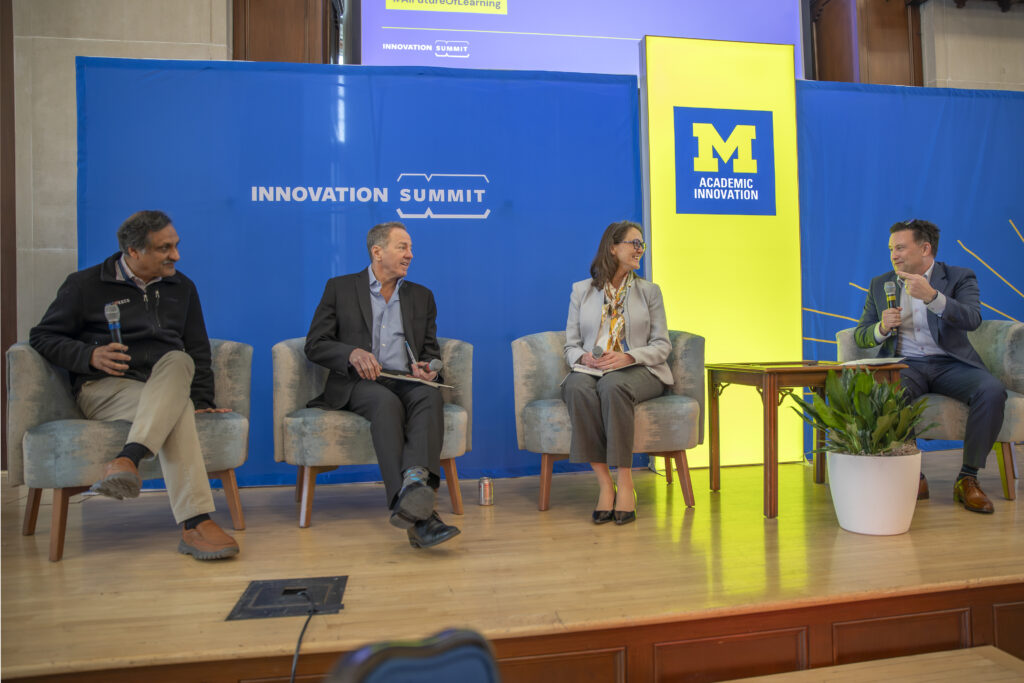Four people sitting side-by-side on a stage, with one person holding a microphone. The background has a block M Academic Innovation sign and another logo on a banner that says Innovation Summit.