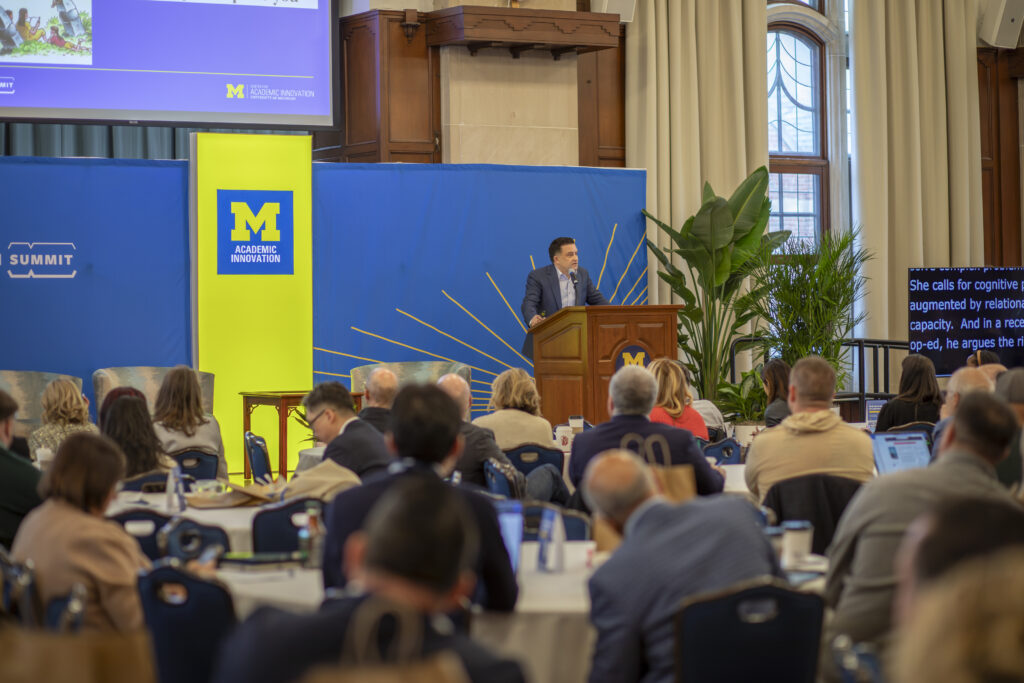 A wideshot of James DeVaney, in a blue suit, speaking to a room full of people in business casual and business professional attire. 