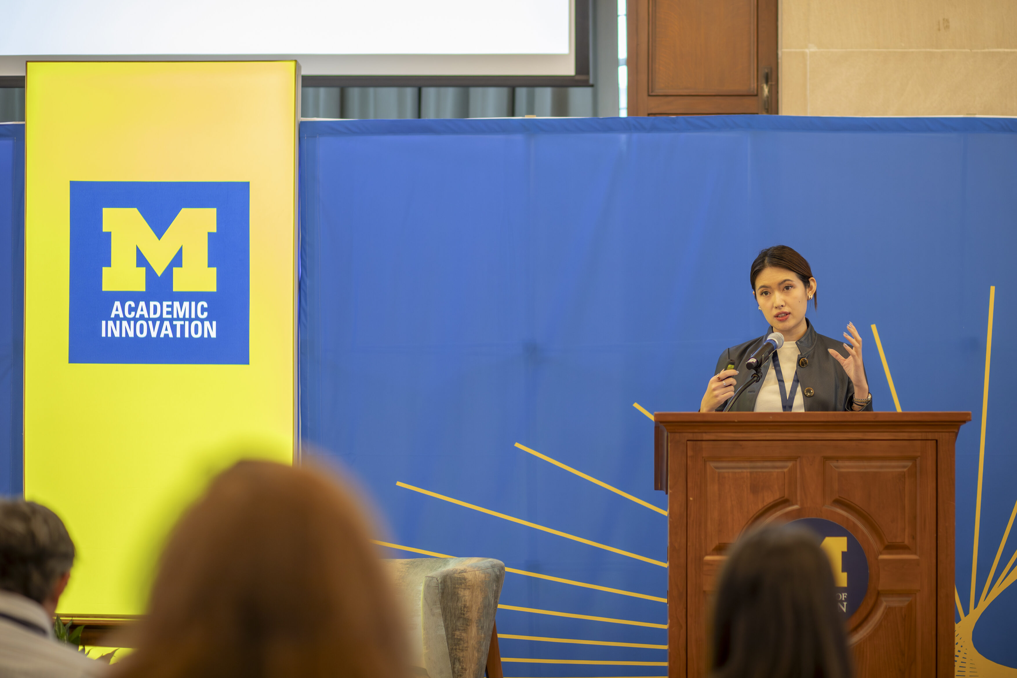Claire Zau stands behind a brown podium, with her hands in the air as she speaks into the microphone. She has dark brownish-black hiar and is wearing a grayish blue jacket over a white T-shirt. 